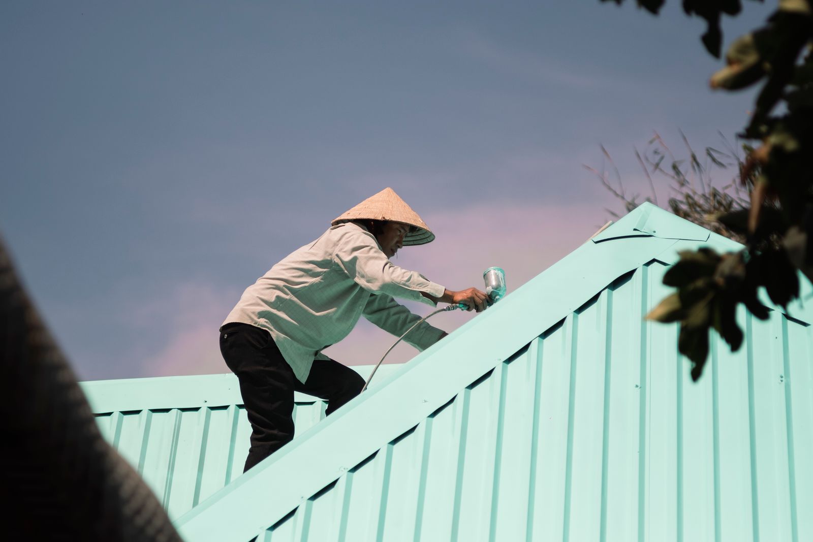 Man in conical hat paints a rooftop, engaging in outdoor work. Low angle view.