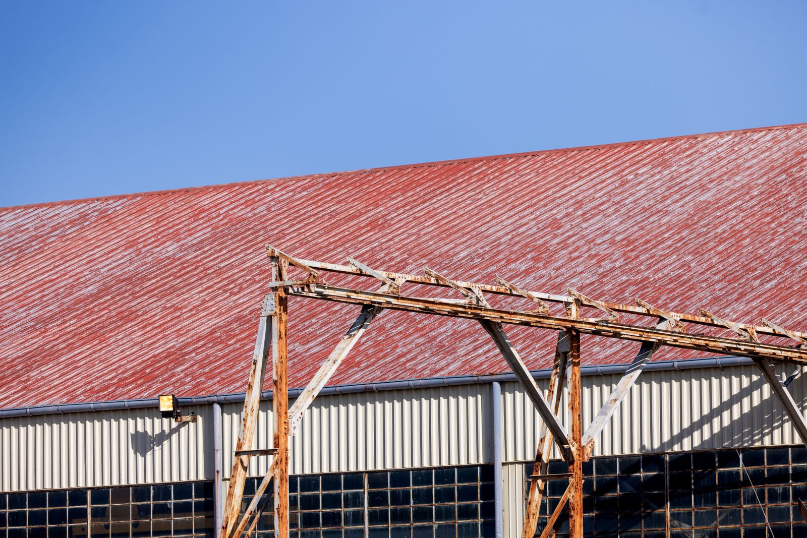 A weathered industrial warehouse with a rusted structure and red roof against a blue sky.