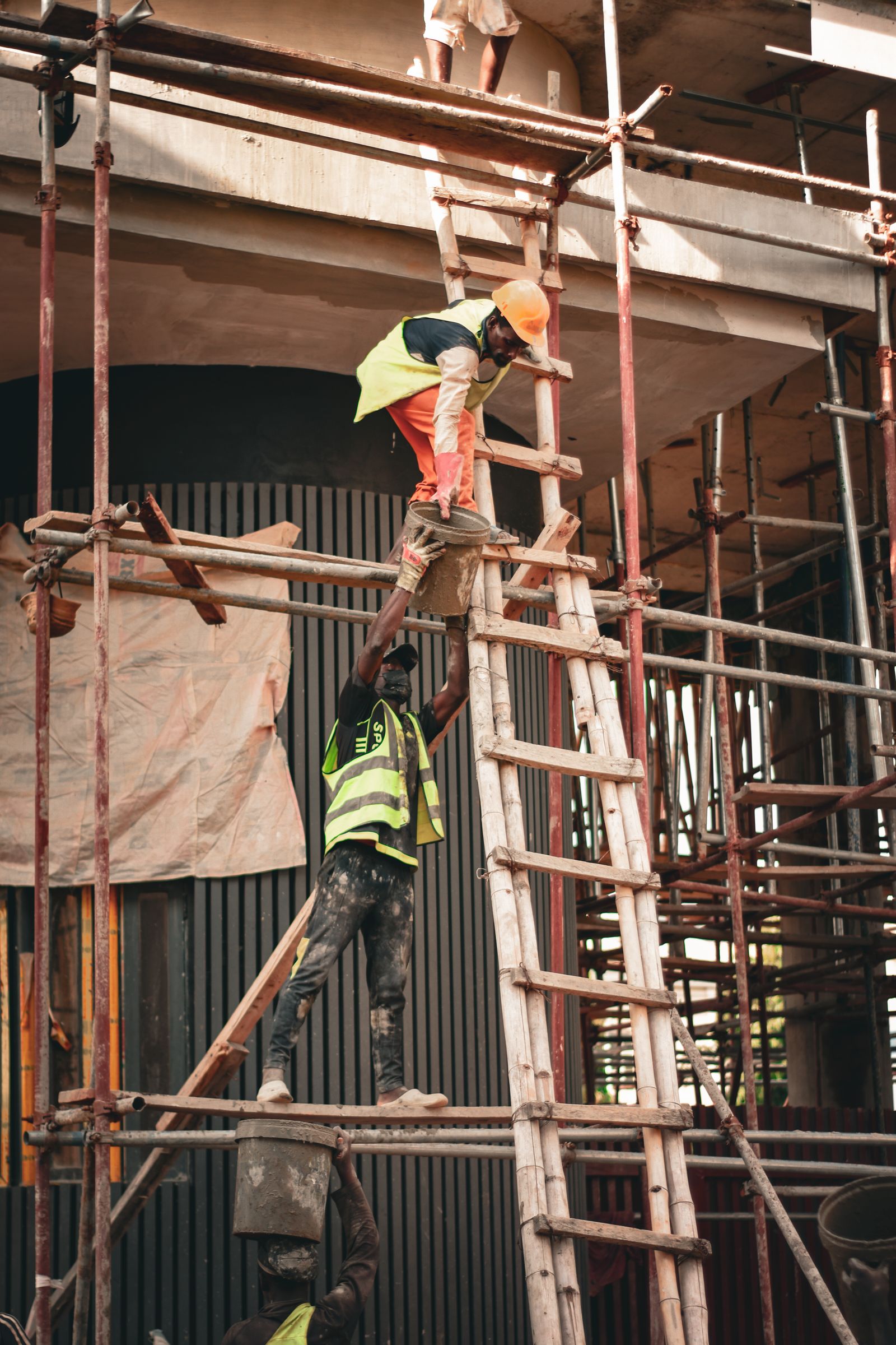 Two construction workers in safety gear collaborate on scaffolding at a building site.